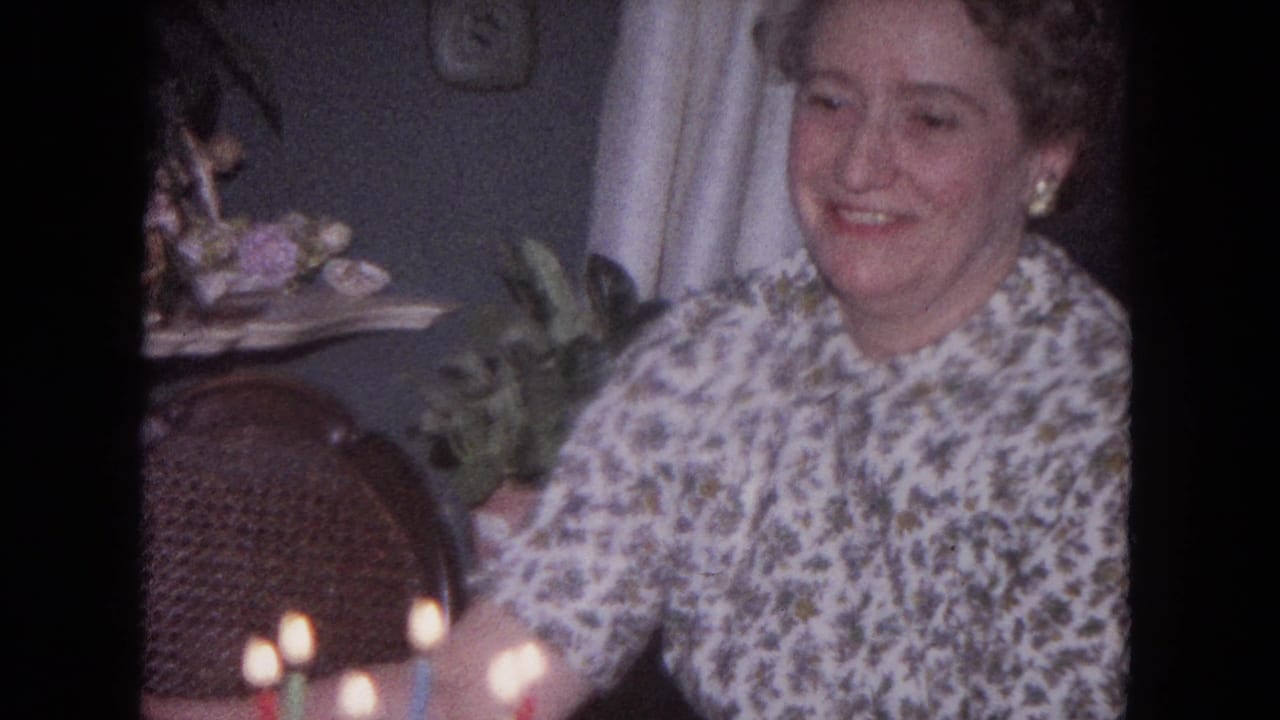 Woman in a floral dress smiles beside lit birthday candles in a dim room.