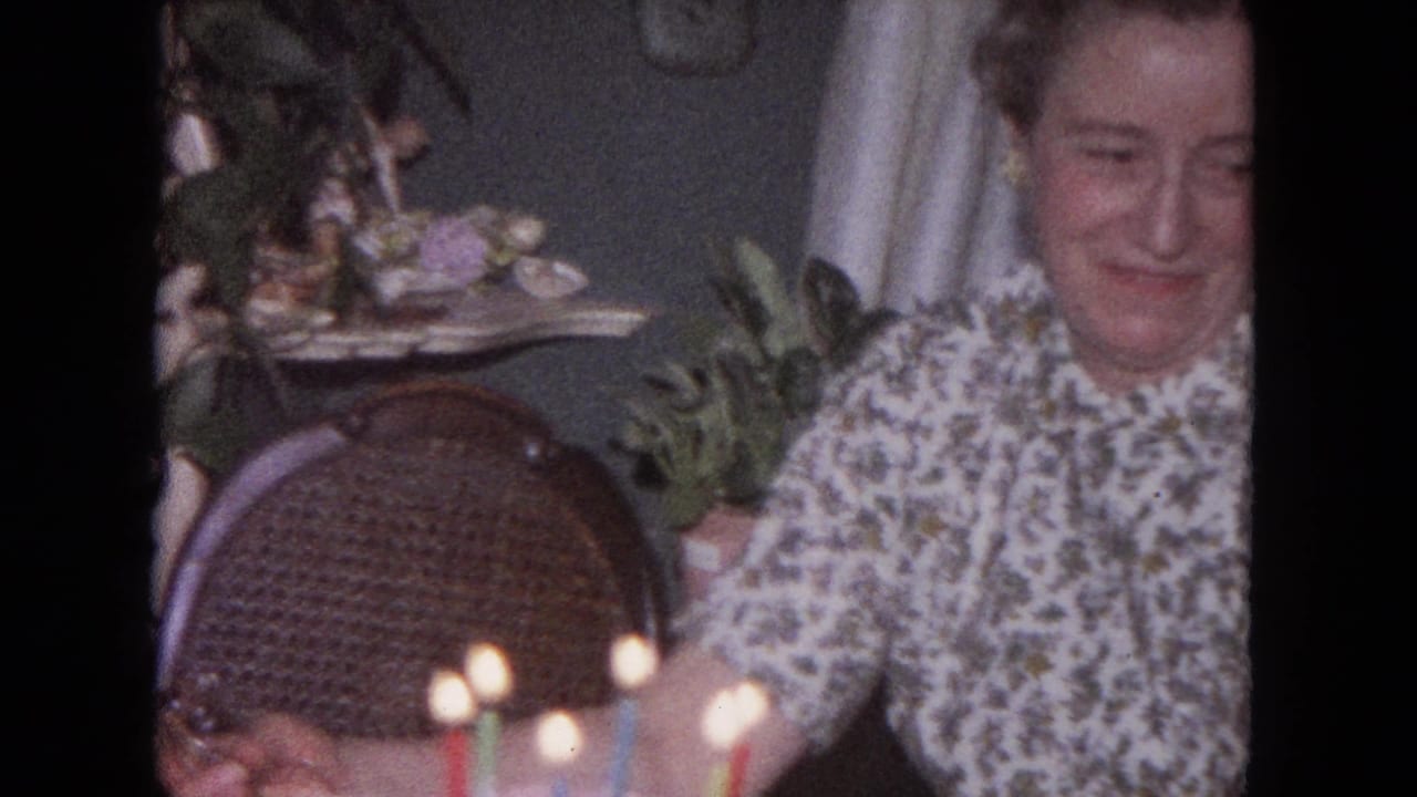 The woman smiles toward the cake and candles, framed by a wicker chair and domestic decor in the 1964 Evanston interior.
