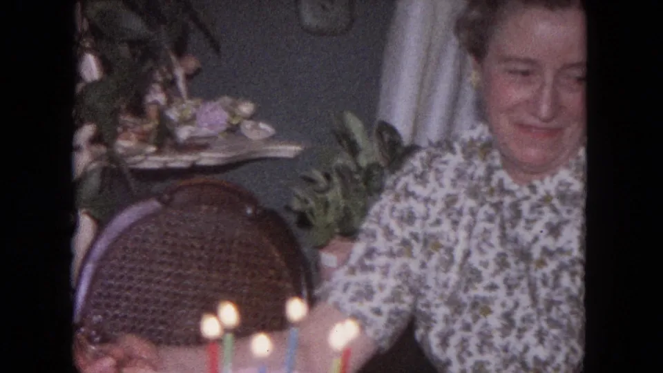 The woman smiles toward the cake and candles, framed by a wicker chair and domestic decor in the 1964 Evanston interior.