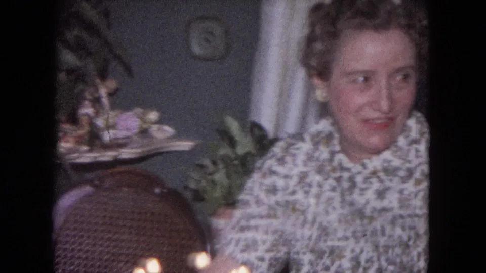 The woman turns sideways near the candlelit cake, with a wicker chair, plant, curtain, and tabletop decorations visible behind her.