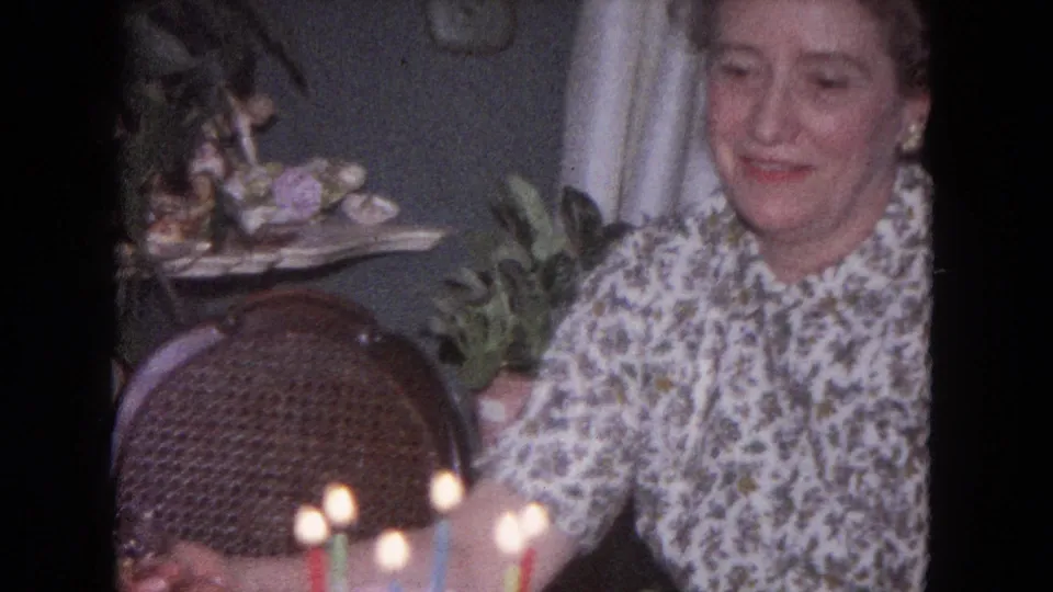 A close home-movie view shows the woman beside the lit birthday cake, with wicker furniture and household decor in the Evanston room.