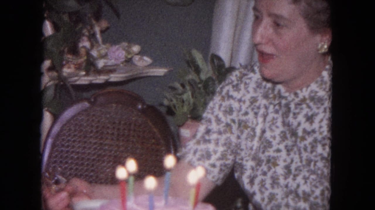 Woman in floral dress seated near a birthday cake with lit candles.