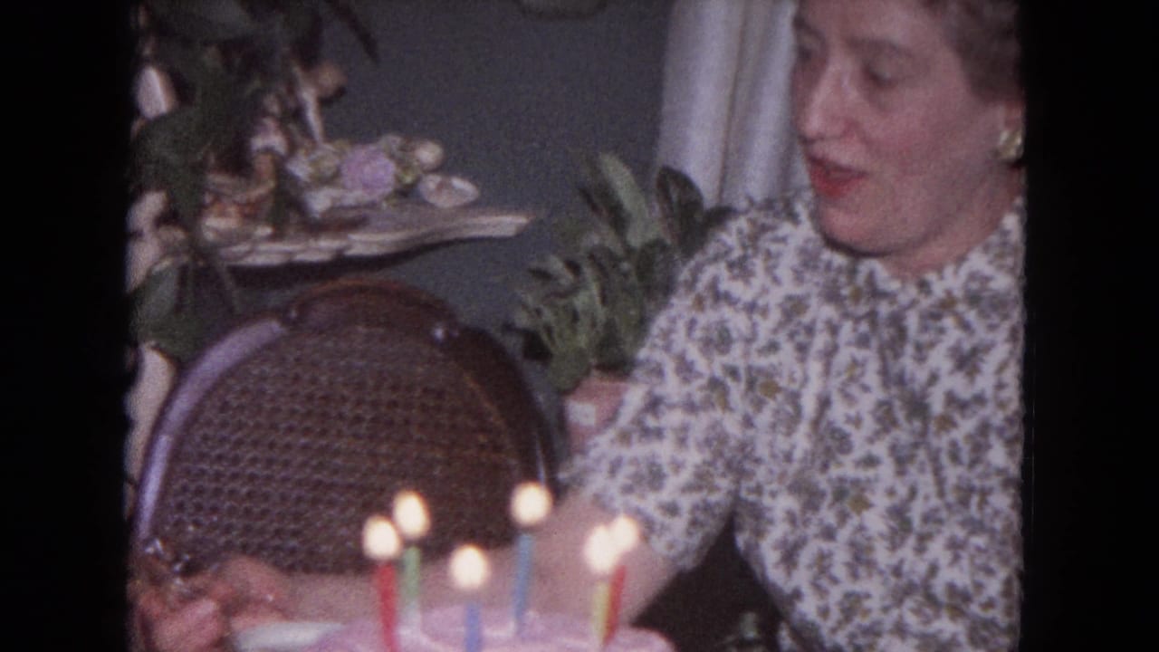 A smiling woman in a floral dress sits near a wicker chair as birthday candles glow in the foreground.