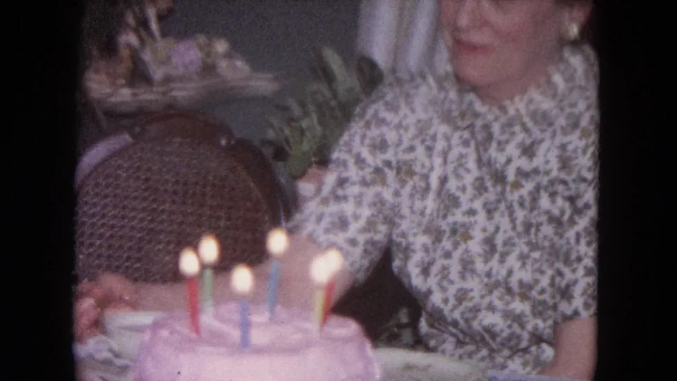 A candlelit birthday cake fills the foreground while the seated woman remains partly visible in the home interior.