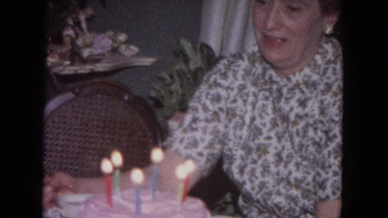 Woman beside a pink birthday cake with lit candles in a dim home interior.
