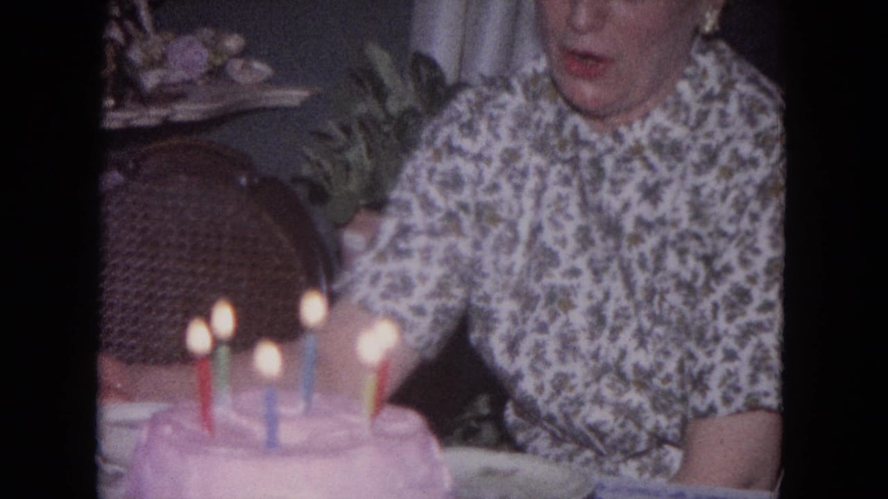 Woman in a floral dress looks toward a birthday cake with several lit candles.