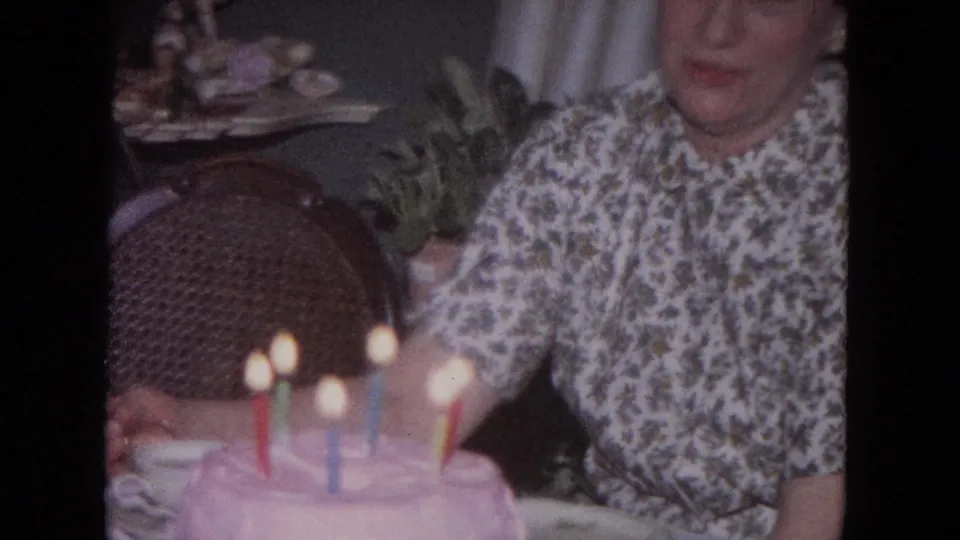 A soft 8mm frame shows the pink frosted birthday cake and candles in front of the seated woman.