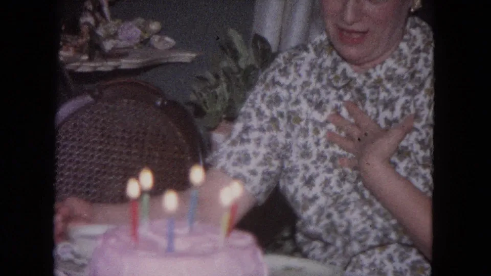 The woman gestures with a hand to her chest near the candlelit cake in the 1964 Evanston birthday scene.