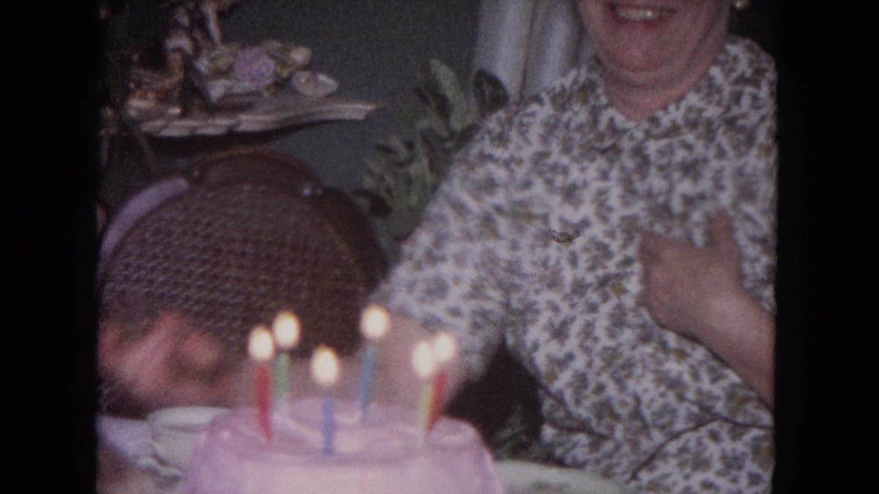 Woman in floral blouse beside a pink birthday cake with lit candles in a home interior.