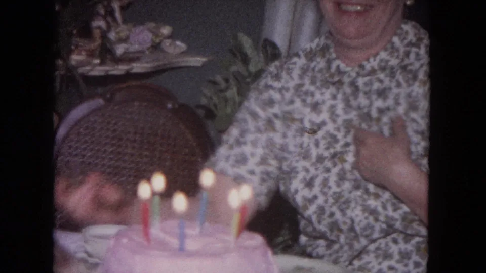 1964 Evanston home-movie frame showing a woman in a floral blouse near a pink birthday cake with lit candles, a wicker chair, and plants.