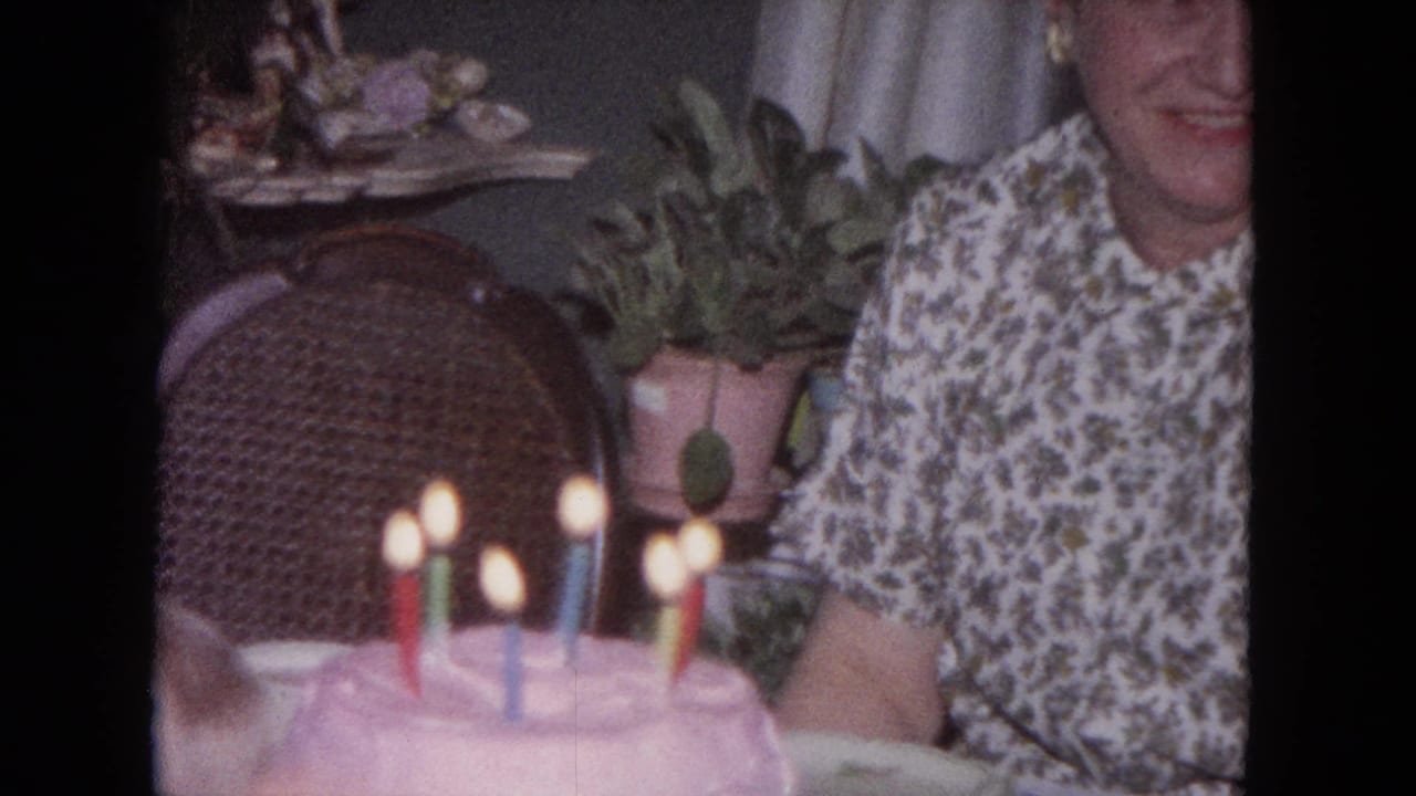 1964 Evanston frame of a smiling woman beside a pink birthday cake with colored candles, with a wicker chair and houseplants in the room.