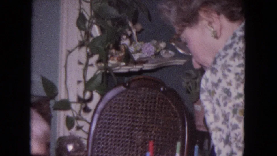 1964 Evanston home interior frame showing the woman leaning near a wicker chair, houseplants, shelf decor, and birthday candles at the lower edge.