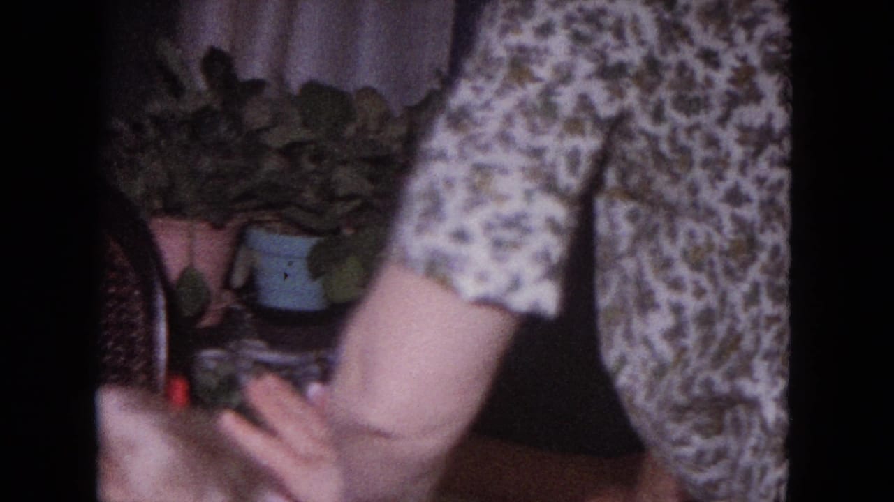 Woman in a floral dress leans near a cake with candles in a home interior.