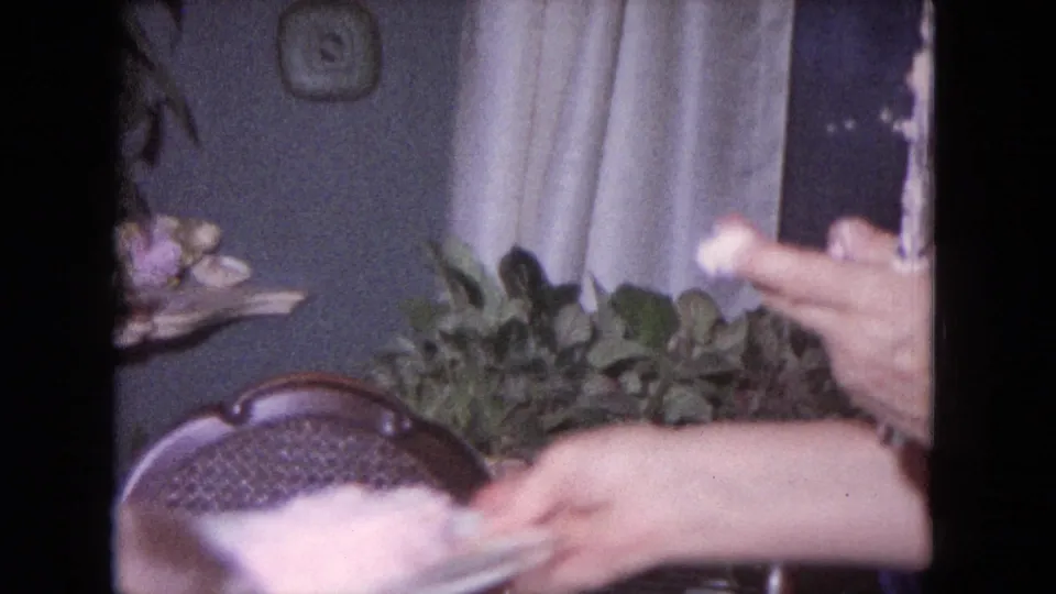 Close home-movie view of hands around a pink-frosted birthday cake as a candle is held near the cake.