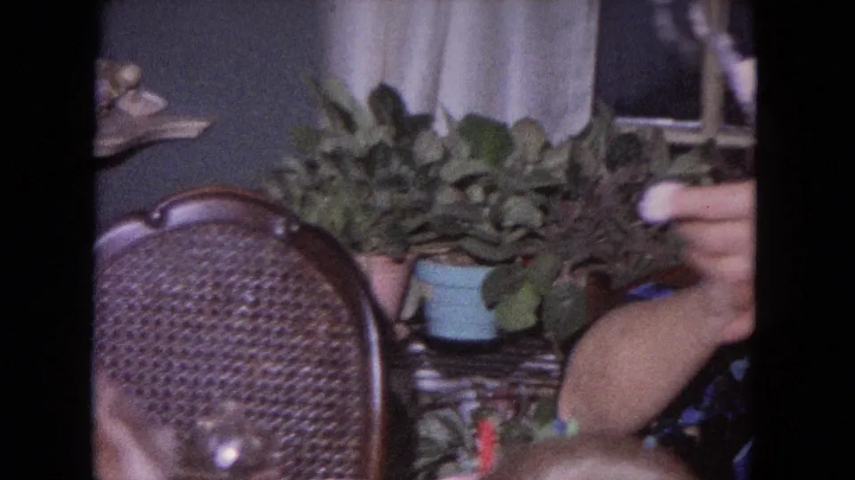 1964 Evanston home interior with wicker chair, potted plant, and hands near the birthday cake as candles are handled.