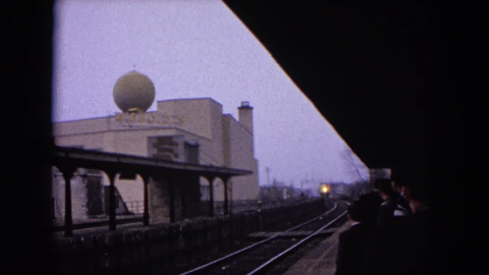 1964 Evanston rail view with tracks, platform canopy, waiting silhouettes, a distant train light, and a building topped by a large sphere.