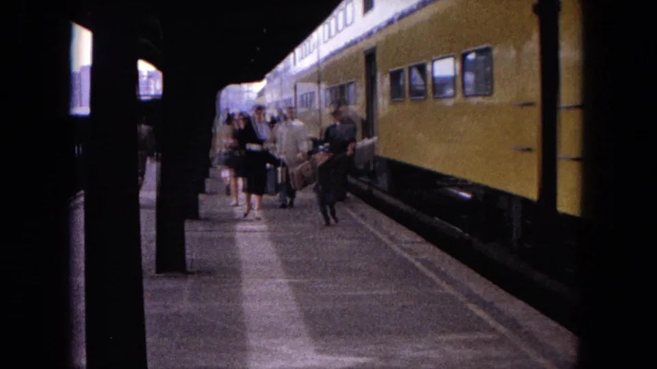Passengers in 1960s dress walk with bags along an Evanston platform beside a stopped yellow train.