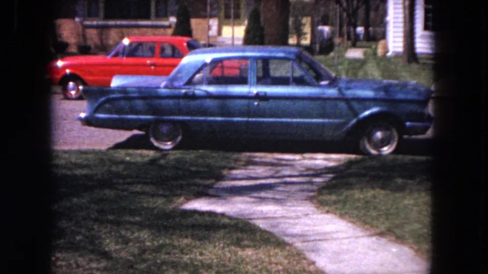 Blue 1960s sedan parked on an Evanston residential street, with a red car behind it and a sidewalk crossing the foreground.