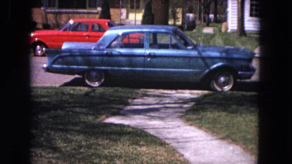 Parked blue sedan and red car on a residential Evanston street, viewed through dark framing in 1964 8mm footage.