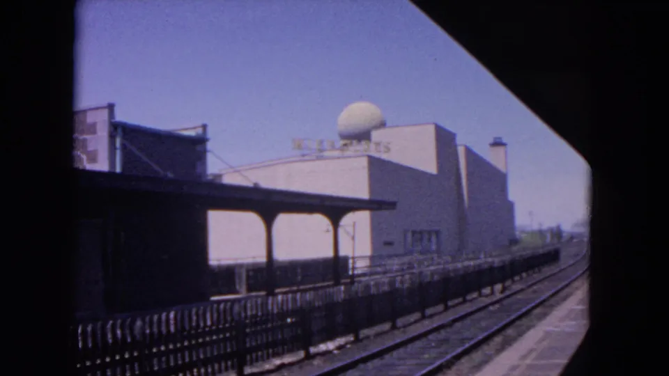 Mid-1960s 8mm view of an Evanston rail platform, dark canopy, fencing, tracks, and Wisconsin signage on a pale building.