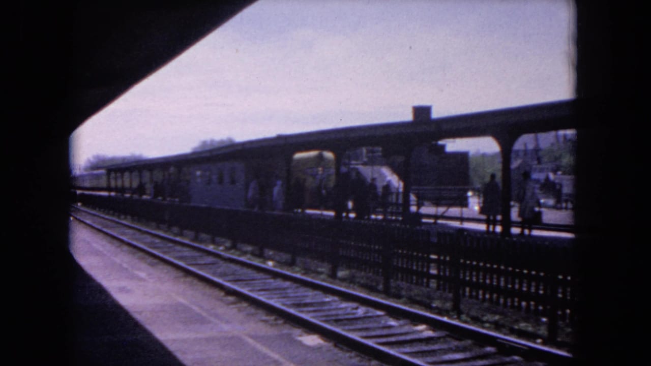 Train tracks lead toward a covered station platform with people waiting in the distance.