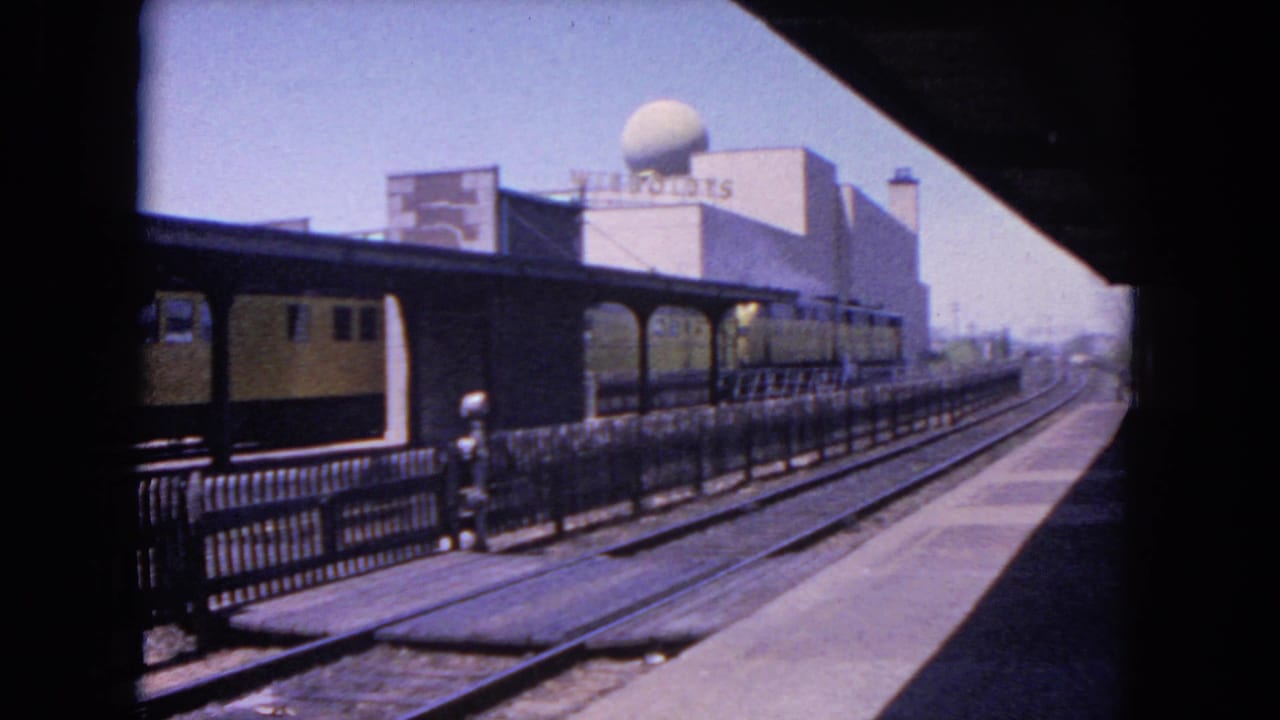 1964 Evanston railway view with the yellow-and-black train moving along the platform, seen from shade with railing, tracks, signage, and a domed building behind.