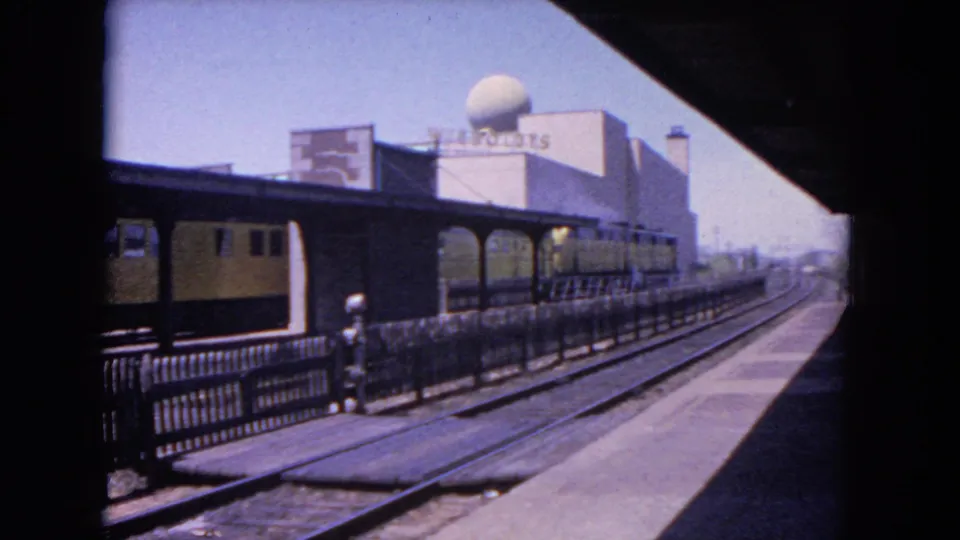 1964 Evanston railway view with the yellow-and-black train moving along the platform, seen from shade with railing, tracks, signage, and a domed building behind.