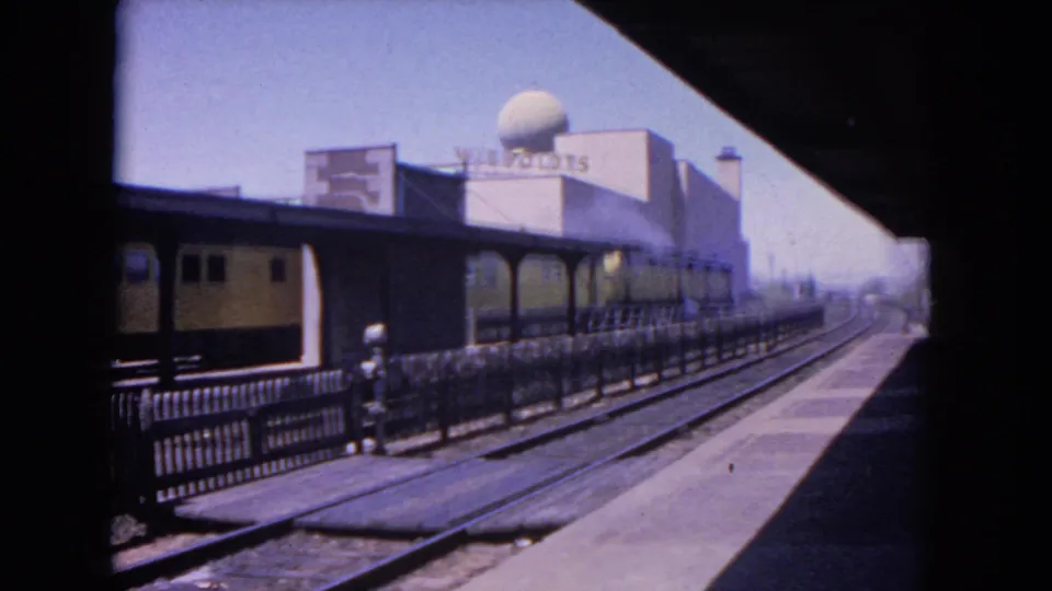 1964 Evanston 8mm frame of a train passing a station platform, with a small figure near the railing and a large domed building visible beyond the tracks.