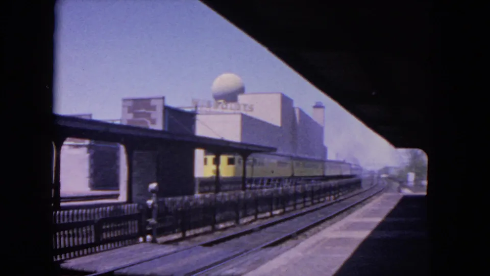 1964 Evanston rail view showing a yellow electric train at a station platform under a canopy, with tracks and a dome-topped building beyond.