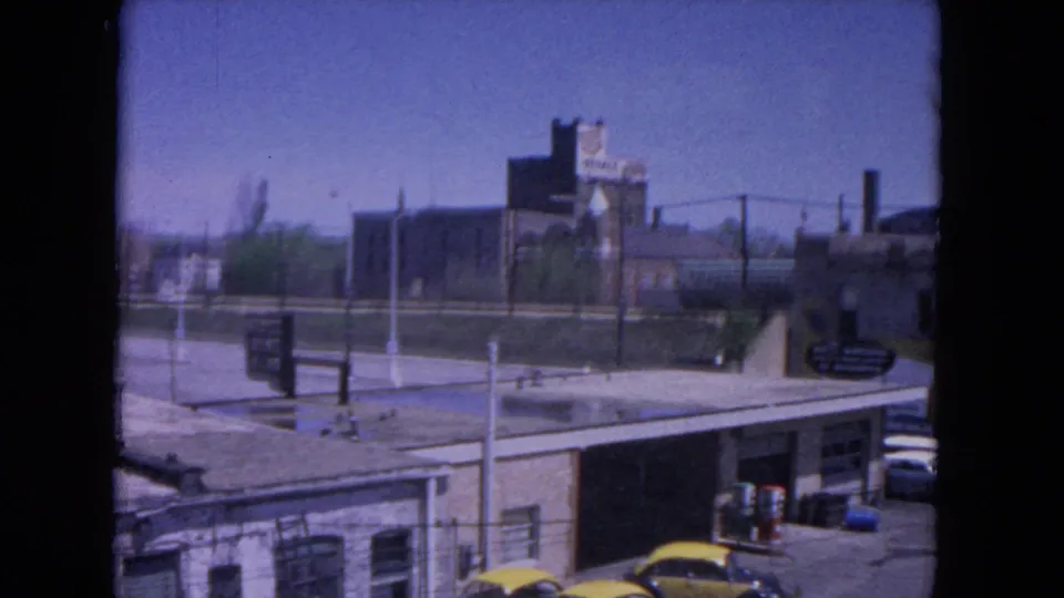 1964 Evanston urban view showing a gas station area, parked cars, signs, utility poles, and industrial buildings near the rail corridor.