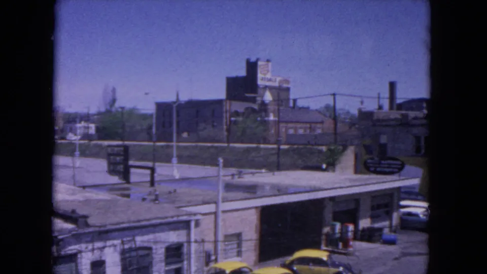 1964 Evanston station-area view with industrial buildings, parked cars, rail platform details, and dark 8mm film edges.
