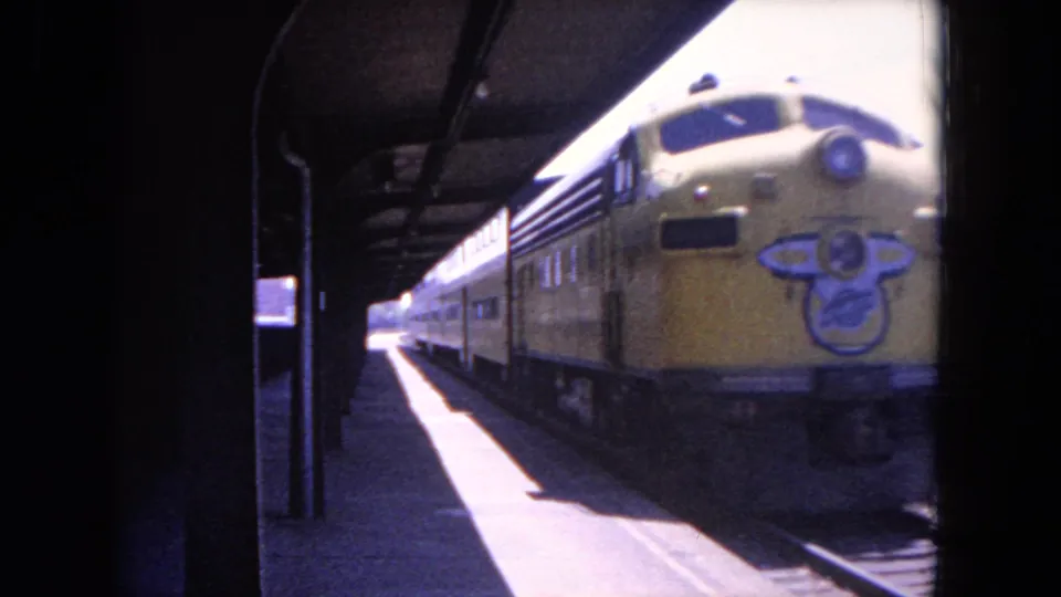 1964 Evanston, Illinois 8mm frame showing a yellow passenger train near a covered station platform with rails and shadows visible.
