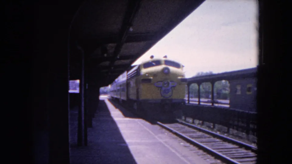 1964 Evanston station view with the yellow train moving along the platform and a darker passenger car visible on the adjacent track.