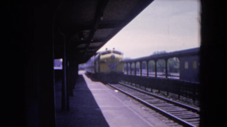 The yellow train appears smaller as it moves away under the Evanston station canopy, with rails and a neighboring car in view.