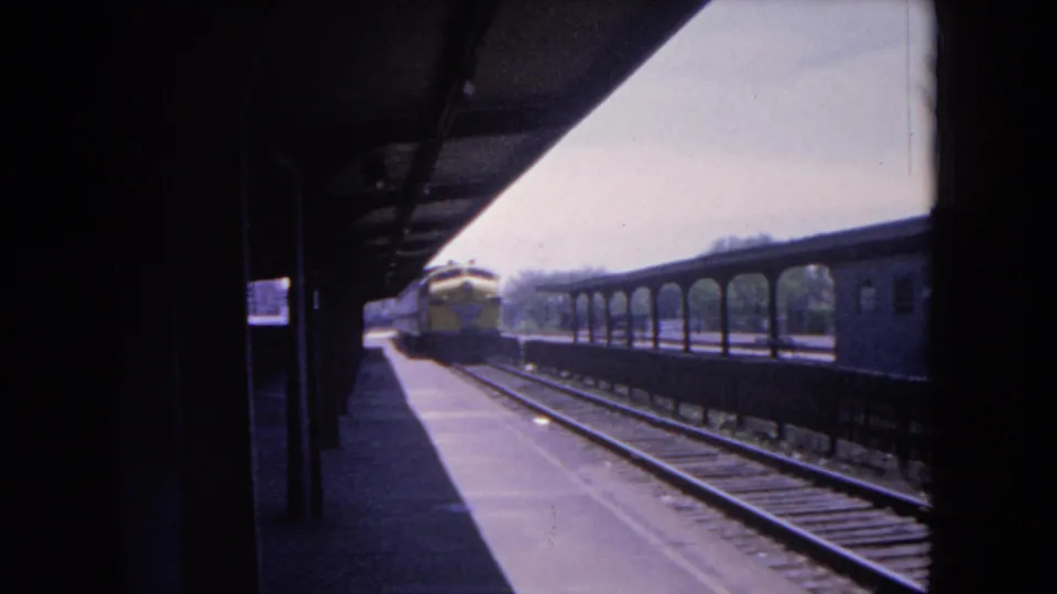 1964 Evanston home-movie frame showing a covered railway platform, tracks, rail cars, and a distant train.