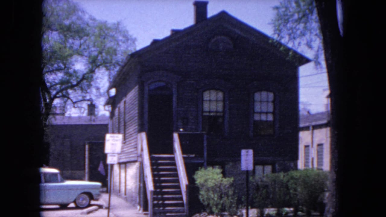 1964 Evanston frame with a dark wood station-area building, arched windows, exterior stairs, signs, shrubs, and a light blue parked sedan.