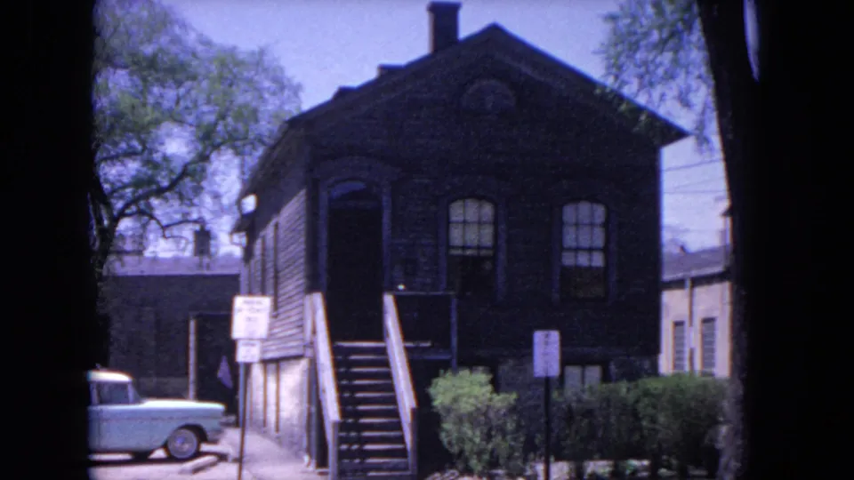 1964 Evanston frame with a dark wood station-area building, arched windows, exterior stairs, signs, shrubs, and a light blue parked sedan.