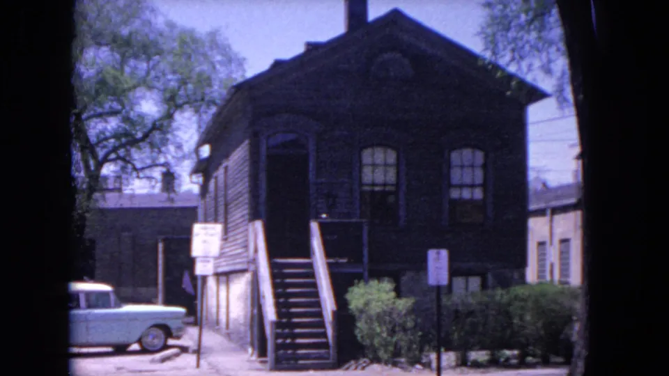 1964 Evanston home-movie view of a wood-sided building with exterior stairs, arched windows, roadside signs, shrubs, and a partially visible blue car.