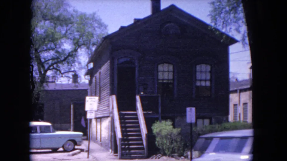 1964 Evanston frame showing the dark wood building, exterior stairs, shrubs, signs, a parked blue sedan, and part of another vehicle in the foreground.
