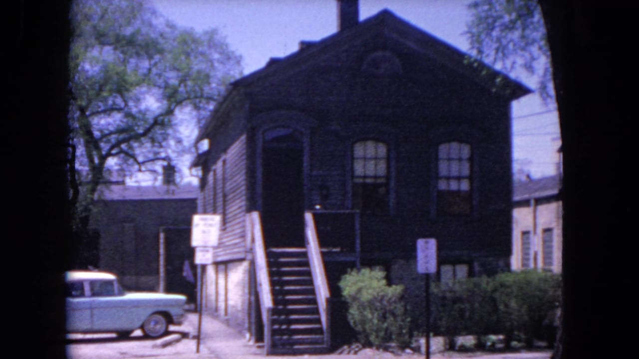 Dark brick building with exterior stairs and a light-colored vintage car parked nearby.