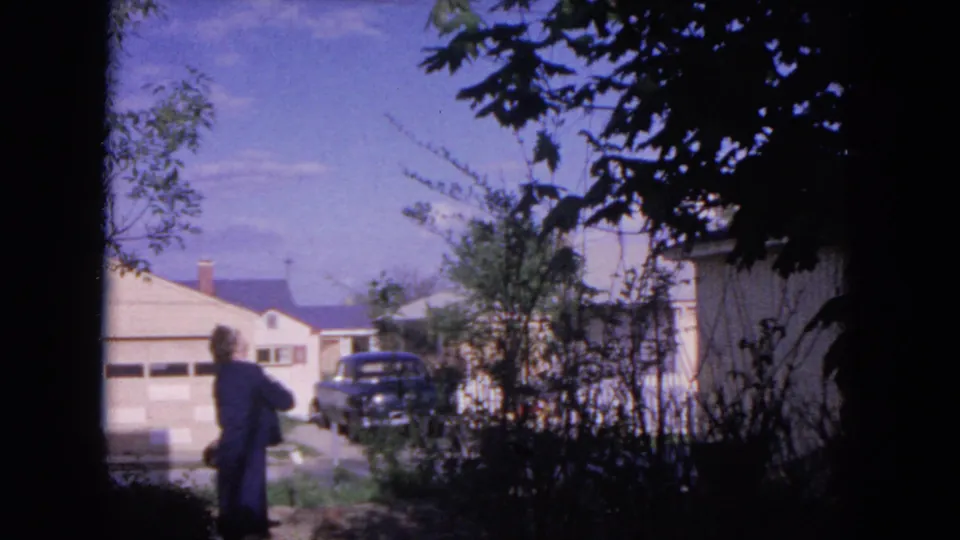 A 1964 Evanston residential street appears with a distant person walking near houses, a garage, a dark car, and overhanging branches.