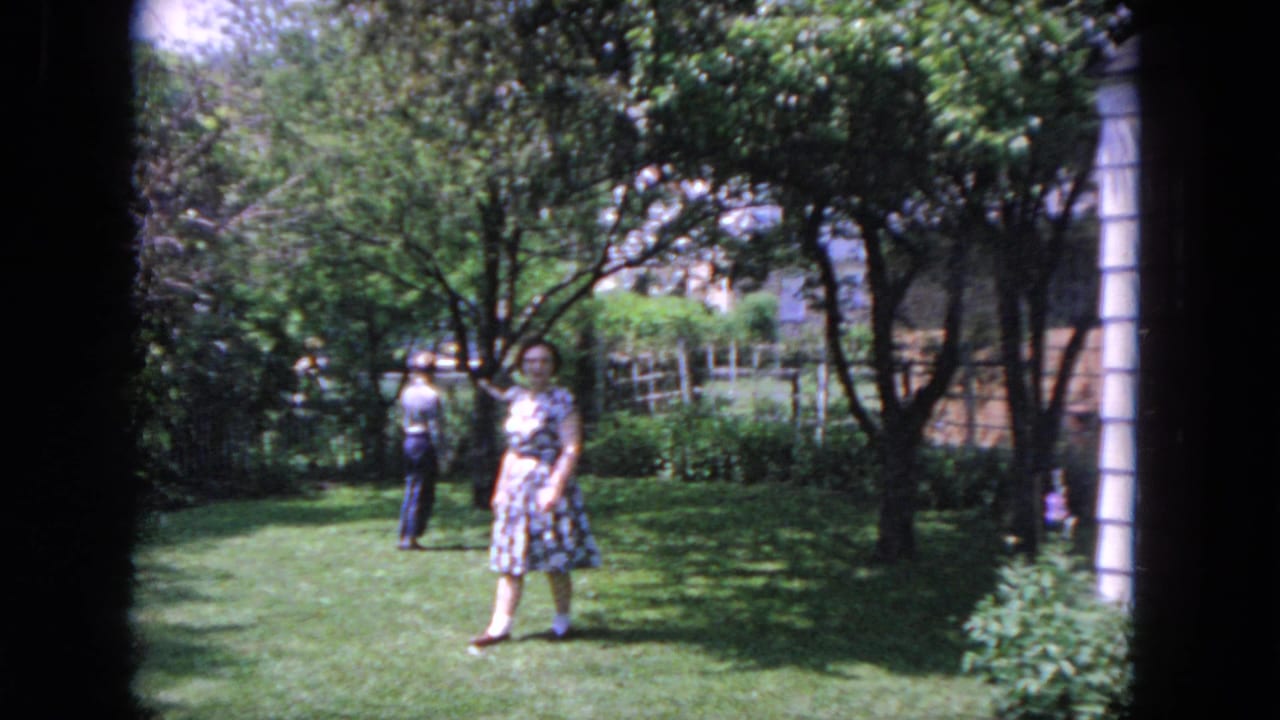 Woman in a patterned dress crossing a sunny backyard lawn with another adult in the background.