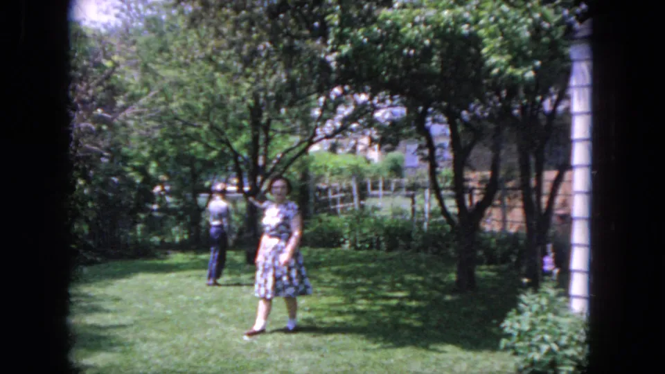 1964 Evanston home-movie frame showing a woman in a patterned dress on a backyard lawn with another adult near trees and fencing.