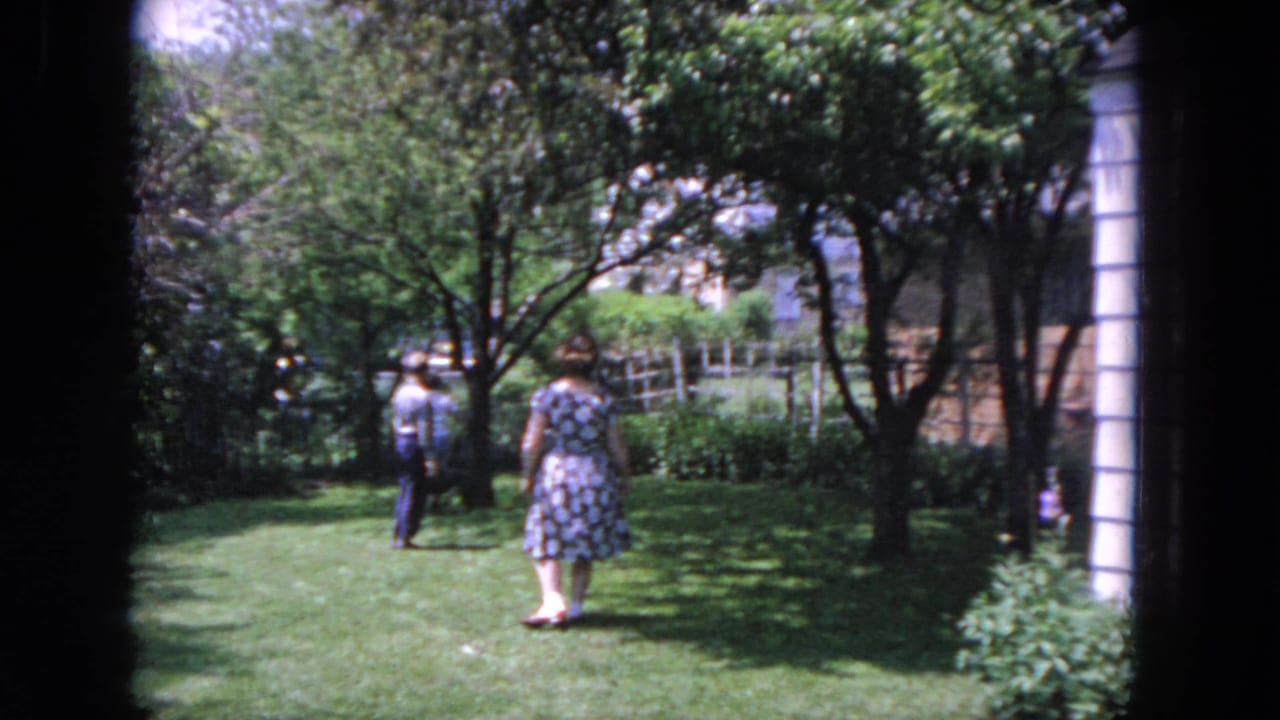 Woman seen from behind walking across the grass toward trees and a fence.