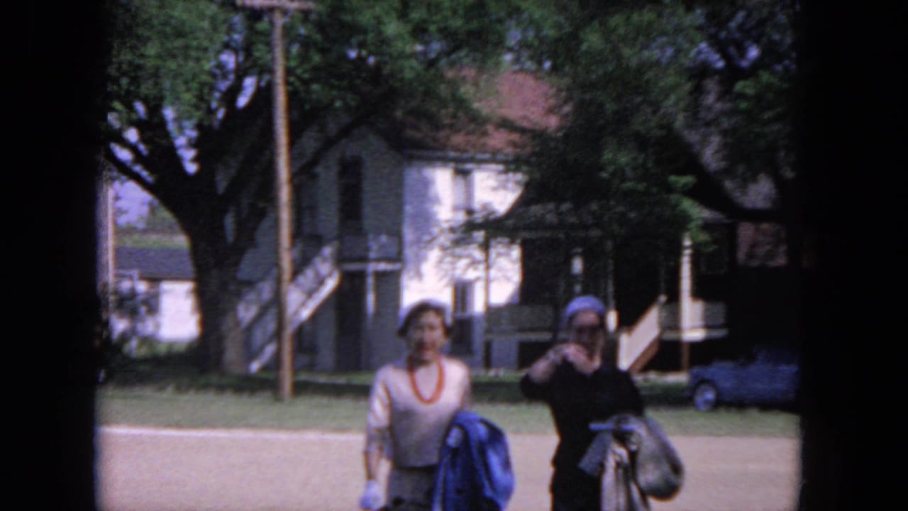 Two dressed women walk across a lawn with a house, trees, and a parked car behind them.