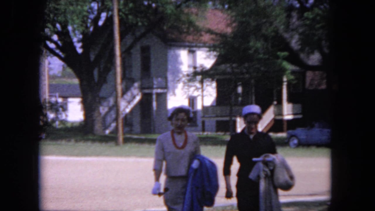 Two women continue walking toward the camera, carrying jackets in front of a house and trees.