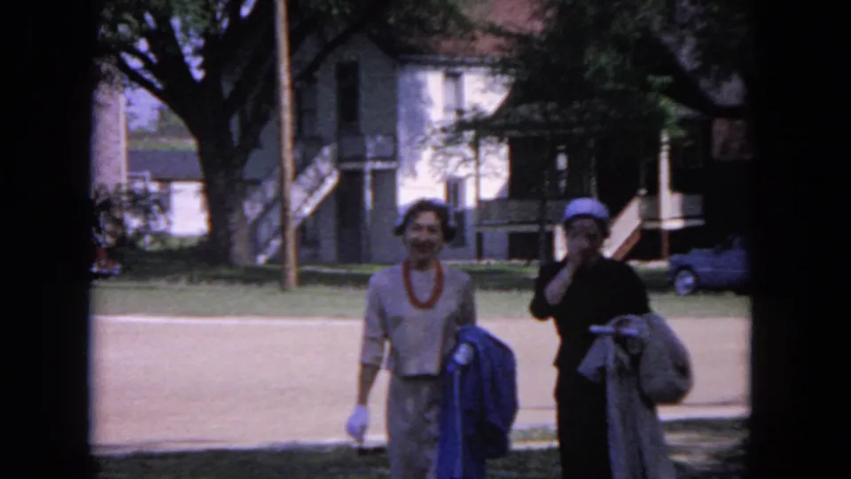 Two women walk side by side in a 1964 Evanston home-movie frame, with trees and residential buildings behind them.