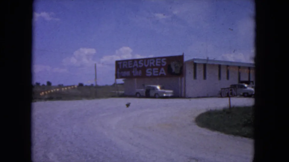 1964 Evanston, Illinois frame showing a low tourist shop building with a Treasures from the Sea sign, parked cars, and roadside entry area.