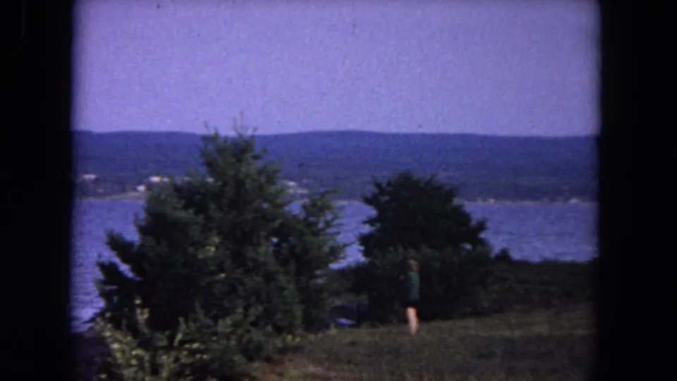 1964 Evanston, Illinois home-movie view of a person on a grassy lakeside overlook, framed by trees with water and distant shoreline beyond.