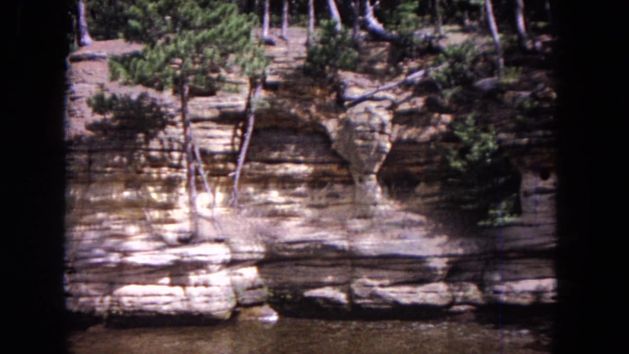 Layered rock bank beside water with small evergreen trees and dark film borders.